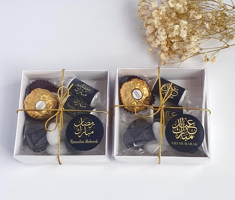 May include: Two white gift boxes with gold ribbon and black and gold labels. The boxes contain chocolate, candy, and a small black gift. The labels on the boxes say "Ramadan Mubarak" and "Eid Mubarak".