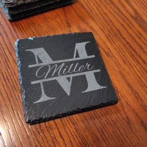 Könnte beinhalten: Schwarzer Schieferuntersetzer mit einem weißen, gravierten Buchstaben "M" und dem Namen "Miller" darunter.