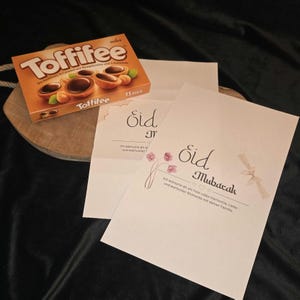 Peut inclure: Une boîte de chocolats Toffifee et deux cartes de vœux avec le texte "Eid Mubarak" en écriture noire. Les cartes ont des motifs floraux et de rubans. La boîte de Toffifee est orange et marron, avec le nom de la marque et la description du produit.