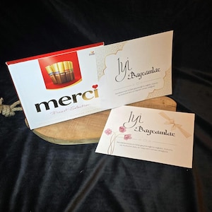 Puede incluir: Una caja de chocolates Merci con un diseño rojo y blanco. La caja tiene un corazón rojo con la palabra "merci" en letras blancas. La caja está sobre una superficie de madera con una tarjeta blanca que dice "İyi Bayramlar" en caligrafía negra. La tarjeta tiene un borde con un patrón dorado y blanco.