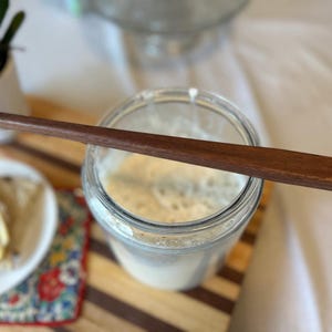 May include: A clear glass jar filled with a light-colored liquid, topped with a dark brown wooden stirring stick. The jar sits on a wooden cutting board. A small white plate with buttered bread is in the foreground.