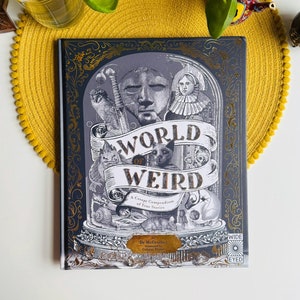Puede incluir: Un libro de tapa dura gris con un diseño dorado y el título "World of Weird: A Creepy Compendium of True Stories" en la portada.