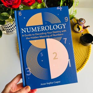 Könnte beinhalten: Ein Hardcover-Buch mit dem Titel "NUMEROLOGIE" mit blauem Einband und goldenen und rosa geometrischen Designs. Der Untertitel des Buches lautet "Ein Leitfaden zur Entschlüsselung Ihres Schicksals mit der verborgenen Bedeutung von Zahlen". Der Name des Autors, Anne-Sophie Casper, steht unten.