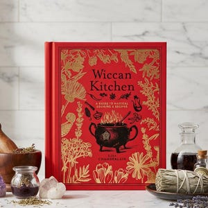 Puede incluir: Un libro rojo de tapa dura titulado "Wiccan Kitchen" con ilustraciones florales doradas y texto. El libro está rodeado de un mortero y una maja, cristales, hierbas y pequeñas botellas. Un gráfico de caldero aparece en la portada.