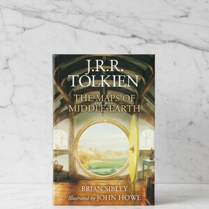 May include: A hardcover book titled "The Maps of Middle-earth" by J.R.R. Tolkien, illustrated by John Howe. The book cover features a detailed illustration of a round doorway with a scenic view, and the author's and illustrator's names.