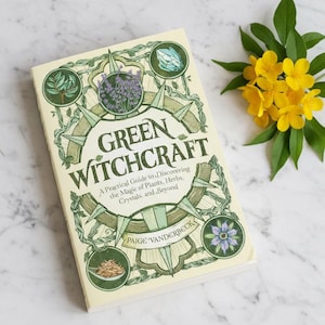 May include: A hardcover book titled "Green Witchcraft" with illustrations of plants and crystals. The book is cream-colored with green and purple accents. A sprig of yellow flowers sits to the right of the book.