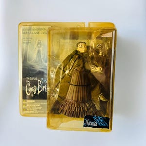 Könnte beinhalten: McFarlane Toys Actionfigur von Victoria aus Tim Burtons Corpse Bride, verpackt in einer transparenten Kunststoffhülle. Die Figur trägt ein braunes Kleid mit Rüschenrock. Die Verpackung enthält den Filmtitel und Victorias Namen.