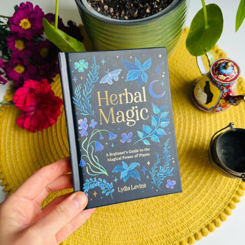 May include: A black book with a blue and green floral design and gold stars. The title is "Herbal Magic: A Beginner's Guide to the Magical Power of Plants" by Lydia Levine.