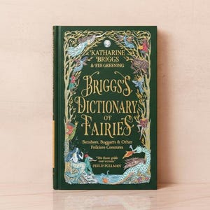 Könnte beinhalten: Ein Hardcover-Buch mit dem Titel "Briggs's Dictionary of Fairies" von Katharine Briggs & Tee Greening. Das Cover ist dunkelgrün mit goldener Schrift und Illustrationen von Feen und Fabelwesen. Das Buch liegt auf einer hellrosa Oberfläche.