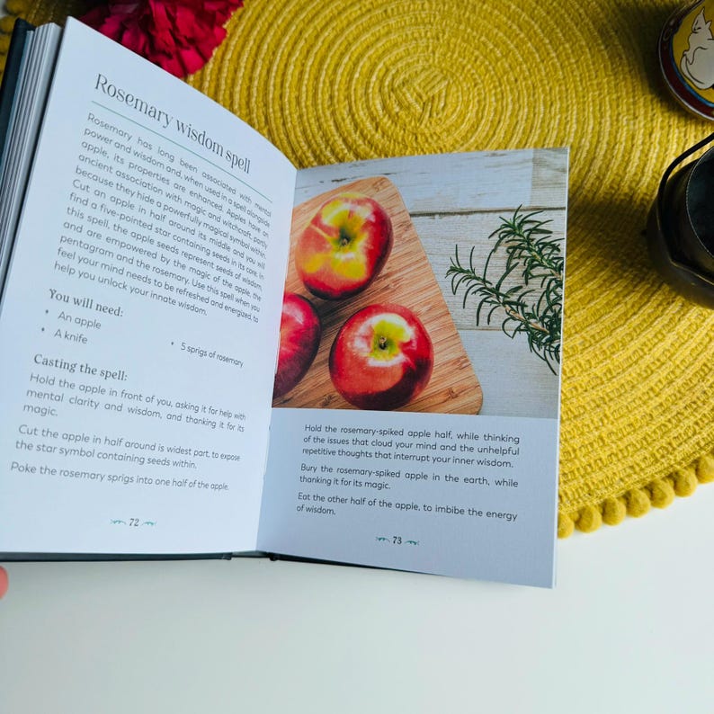 May include: Open book with a white background showing a spell for wisdom using rosemary and an apple. The book has a title "Rosemary Wisdom Spell" and instructions on how to perform the spell. The image shows two red apples on a wooden cutting board with a sprig of rosemary.