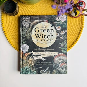 May include: A hardcover book titled "The Green Witch Illustrated" with botanical illustrations and the text "An Enchanting Immersion Into The Magic Of Natural Witchcraft." The book is on a yellow placemat with a decorative edge, with a small cup and flowers in the background.