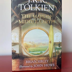 May include: A book titled "The Maps of Middle-earth" by J.R.R. Tolkien, illustrated by John Howe. The cover features a detailed illustration of a round doorway overlooking a landscape. The book is on a red surface.