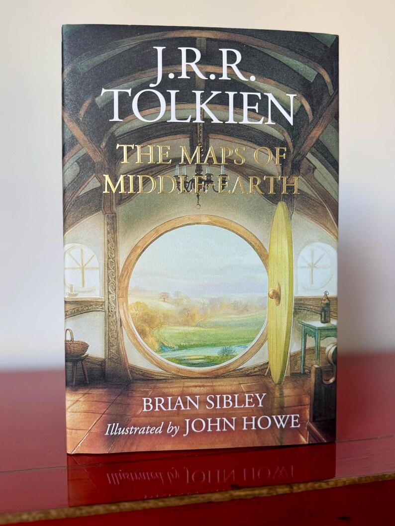 May include: A book titled "The Maps of Middle-earth" by J.R.R. Tolkien, illustrated by John Howe. The cover features a detailed illustration of a round doorway overlooking a landscape. The book is on a red surface.