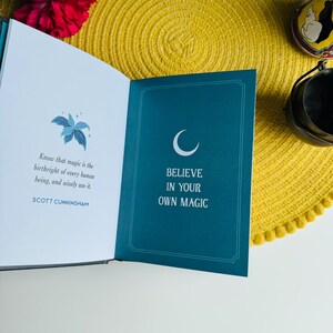 May include: Open book with a blue cover and white text that reads "Believe in your own magic". The book is open to a page with a crescent moon illustration and a quote by Scott Cunningham that reads "Know that magic is the birthright of every human being, and wisely use it."