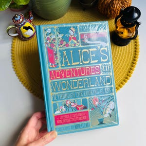 Puede incluir: Un libro de tapa dura titulado "Alice's Adventures in Wonderland & Through the Looking-Glass". El libro tiene una cubierta turquesa con letras e ilustraciones doradas. Ilustrado por Minalima.