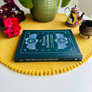 May include: A dark green hardcover book titled "The Wiccan Handbook: A modern guide to the symbols, spells and rituals of witchcraft" by Susan Bowes. The book is sitting on a yellow circular placemat.