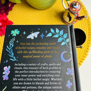 May include: A black book with a gold title that reads "Step into the enchanting world of herbal recipes, remedies and rituals with this spellbinding guide to the magical power of plants". The book cover features a crescent moon, stars, butterflies, and green leaves.