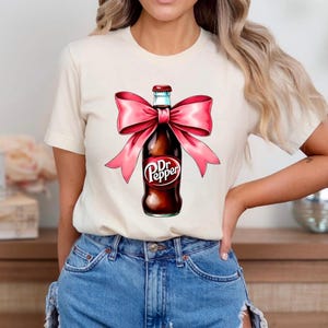 May include: A white t-shirt with a pink bow tied around a brown glass bottle with the text "Dr Pepper" on the label.