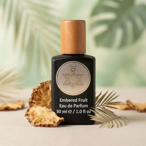 Puede incluir: Un frasco de perfume negro con tapón de madera, con un emblema dorado con una estrella y una luna creciente. La etiqueta dice "Embered Fruit Eau de Parfum 30 ml". Frutas secas y follaje rodean el frasco.