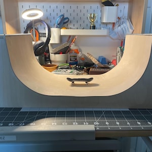 Puede incluir: Una rampa de half-pipe en miniatura de madera con una pequeña tabla de skate en la parte superior. La rampa está hecha de madera contrachapada y tiene un acabado liso de color marrón claro. La tabla de skate es negra y tiene ejes y ruedas plateados. La rampa está colocada sobre una estera de corte negra con marcas blancas. La estera de corte está sobre una mesa de madera con un borde blanco.