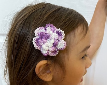 May include: A purple and white crocheted flower hair clip. The flower is made of multiple layers of crocheted petals.
