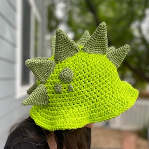 May include: A bright green crochet hat, designed to resemble a dinosaur. The hat has a series of green spikes along the top and a rounded crown. The spikes are a darker shade of green than the hat's main body.