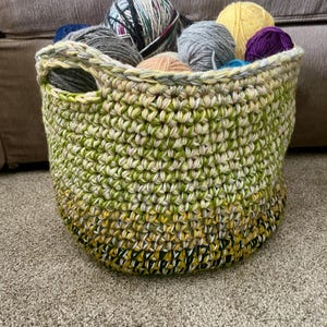May include: A handmade crochet basket in shades of green, yellow, and cream, filled with various yarn balls in colors like gray, purple, and yellow. The basket has two handle openings and sits on a neutral-colored carpet.