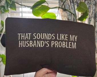 Może przedstawiać: Ciemnobrązowa bluza z haftowanym białym napisem "THAT SOUNDS LIKE MY HUSBAND'S PROBLEM". Bluza jest trzymana przed oknem z roślinami i zasłonami w tle.