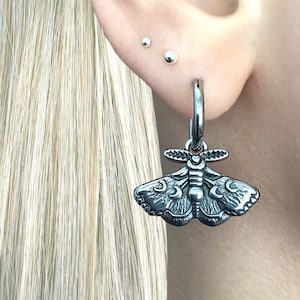 May include: A silver hoop earring with a detailed moth pendant. The moth design features intricate wing patterns and body details. The earring is attached to a silver hoop, creating a unique and stylish accessory.