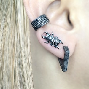 May include: A pair of silver earrings, one featuring a beetle design and the other a geometric shape. The earrings are worn in the earlobe and helix piercing.