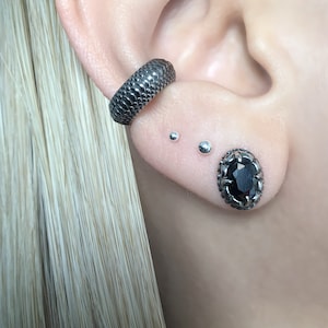 May include: A close-up of a person's ear with multiple piercings. The ear is pierced with a black textured hoop earring, two small silver stud earrings, and a larger black oval stud earring with a silver setting.