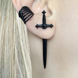 May include: Black sword-shaped earring with a matching ear cuff. The earring features a detailed hilt with a central gem and cross-shaped accents. The ear cuff has a layered design. The jewelry is worn on a person's ear.