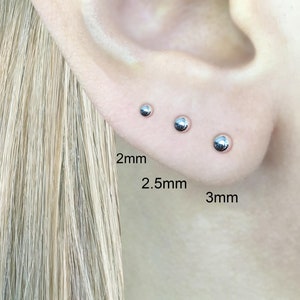 以下が含まれることがあります： 2mm、2.5mm、3mmの3つの異なるサイズのシルバーボールスタッドピアス。ピアスは人の耳につけられています。