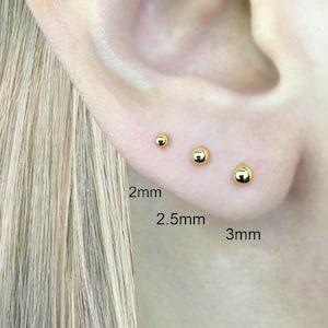Puede incluir: Tres pendientes de bola de oro en diferentes tamaños: 2 mm, 2,5 mm y 3 mm. Los pendientes se muestran en el lóbulo de la oreja de una persona.