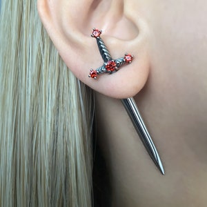 May include: A silver sword earring with red gemstones. The sword is detailed with a textured design and has a long, pointed tip.