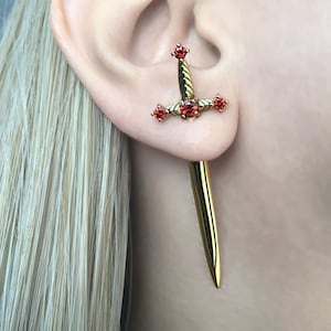 以下が含まれることがあります： 赤い宝石が付いた金の剣のイヤリング。剣は長く、先が尖った細長い形をしています。イヤリングは耳たぶにつけられています。
