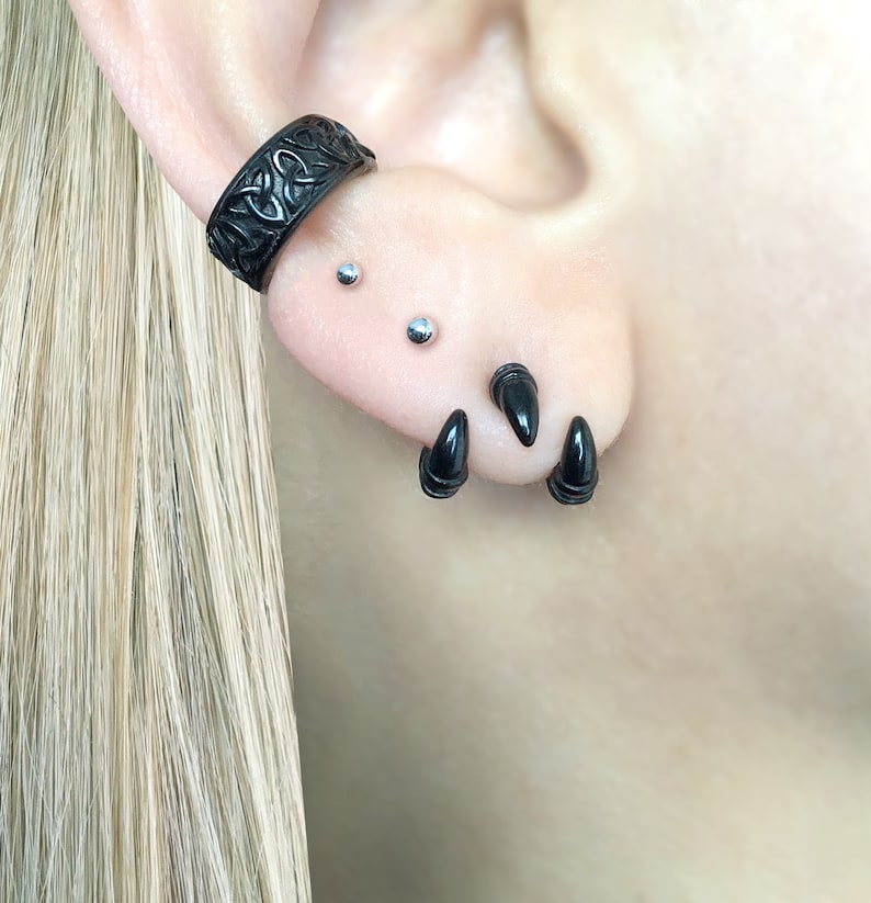 May include: Black ear cuff with a Celtic knot design, and three black spike earrings. The earrings are worn in the upper ear lobe and helix.