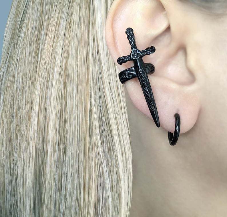 May include: Black ear cuff with a sword design and a black hoop earring. The jewelry is worn on the left ear.