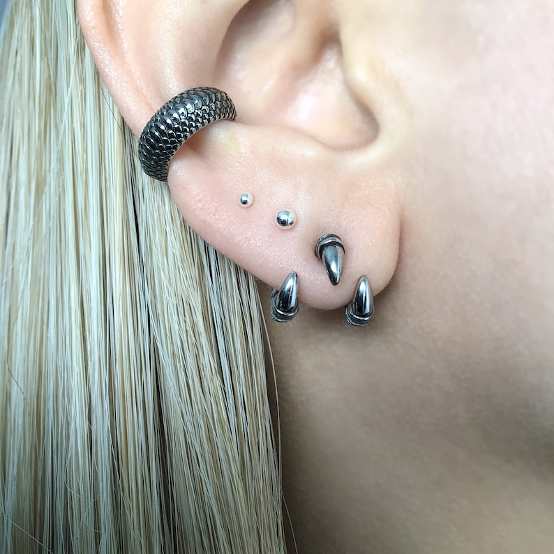 May include: A close-up of a person's ear with multiple piercings. The ear is pierced with a silver hoop earring, two small silver stud earrings, and three silver spike earrings.