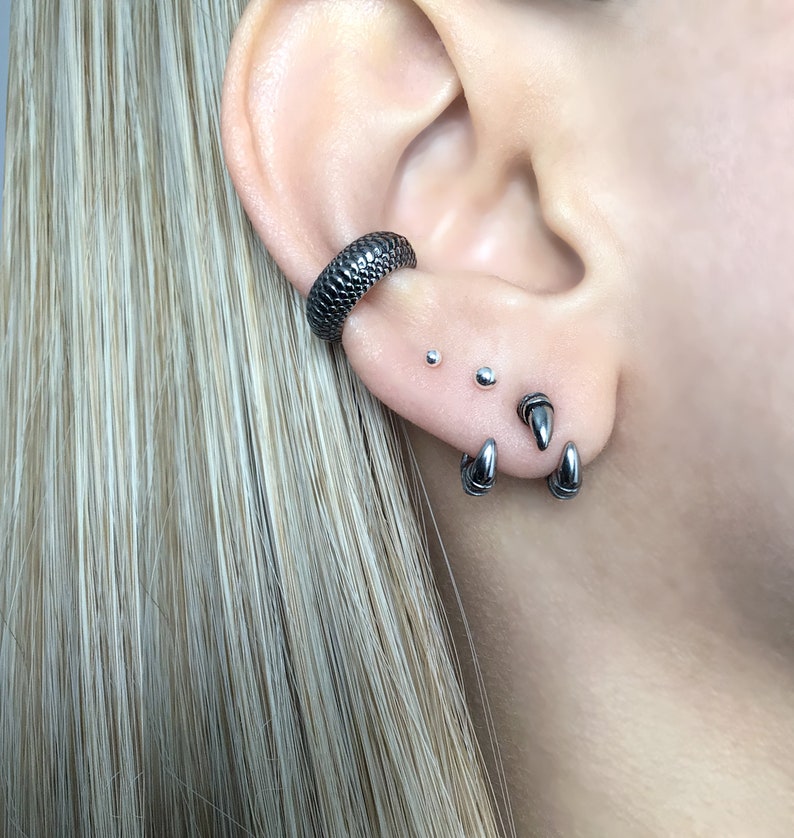 May include: A close-up of a person's ear with multiple silver earrings. The earrings include a small hoop, two small studs, and a pair of spike earrings.