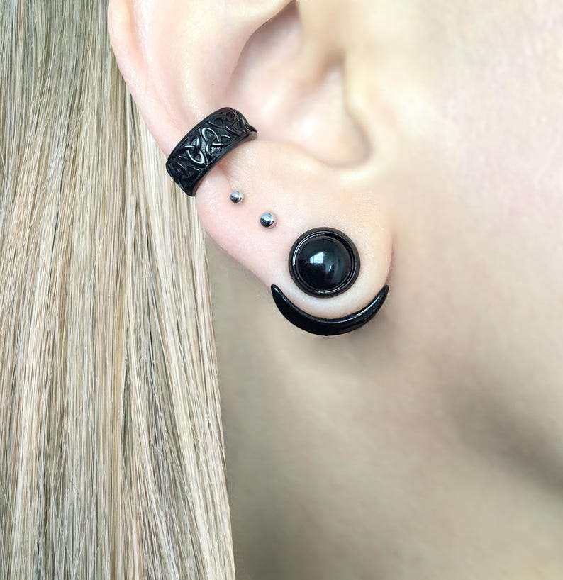 Puede incluir: Un brazalete de oreja negro, un pendiente de media luna negro y un tap&oacute;n de calibre redondo negro con una piedra negra en el centro.