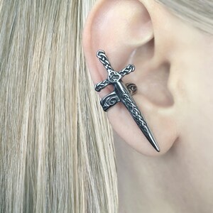 May include: A silver ear cuff in the shape of a sword with intricate Celtic knot designs. The cuff is worn on the upper part of the ear.