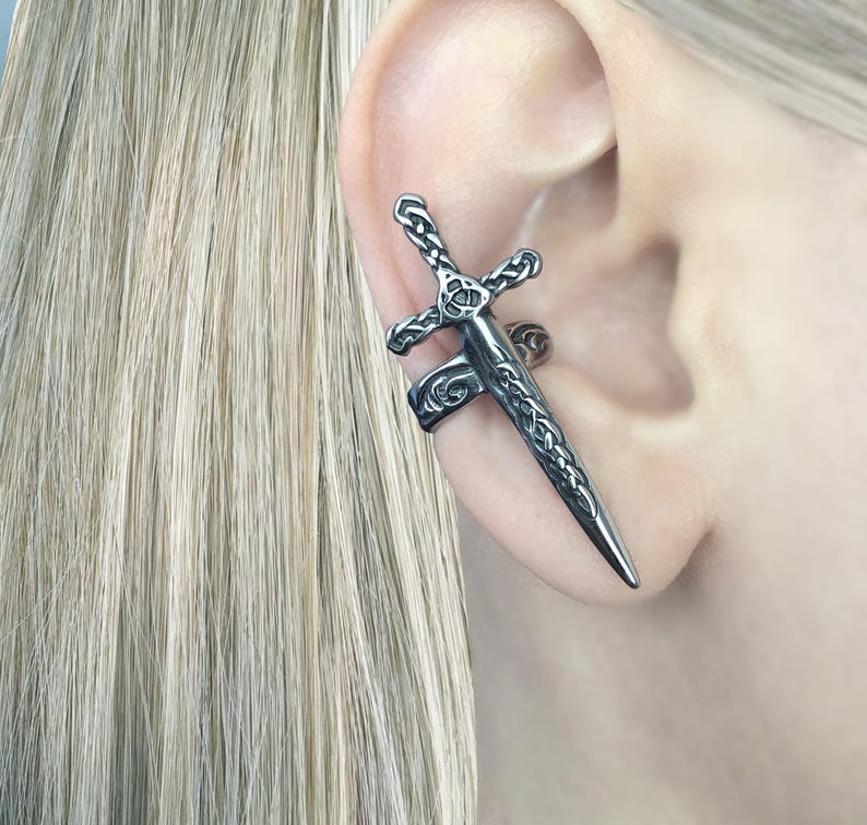 May include: A silver ear cuff in the shape of a sword with intricate Celtic knot designs. The cuff is worn on the upper part of the ear.