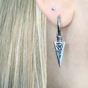 May include: A silver-toned hoop earring featuring a dangling pendant shaped like a pointed arrowhead. The pendant is decorated with an intricate, raised Celtic triquetra knot pattern and has a polished metallic finish with darkened recessed details.