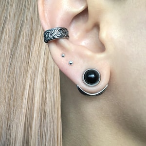 Puede incluir: Una oreja con m&uacute;ltiples piercings. La oreja est&aacute; perforada con un pendiente de aro de plata con un dise&ntilde;o de nudo celta, dos peque&ntilde;os pendientes de plata y un aro de plata con una piedra negra.