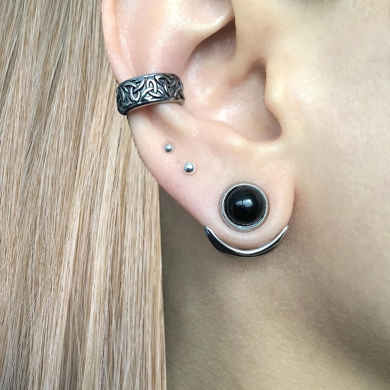 Puede incluir: Una oreja con m&uacute;ltiples piercings. La oreja est&aacute; perforada con un pendiente de aro de plata con un dise&ntilde;o de nudo celta, dos peque&ntilde;os pendientes de plata y un aro de plata con una piedra negra.