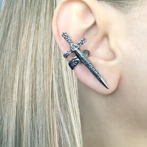 May include: A silver sword ear cuff with intricate detailing. The sword is wrapped around the ear and has a pointed tip.