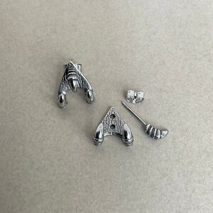 May include: A pair of silver-toned earrings with a spaceship design. The earrings are made of metal and have a textured surface. The earrings are shown on a light brown surface.