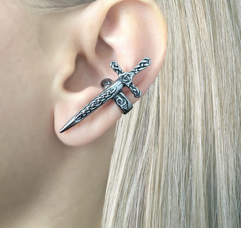 May include: A silver ear cuff in the shape of a sword with intricate details. The sword is positioned vertically with the point facing down. The ear cuff is designed to wrap around the ear.