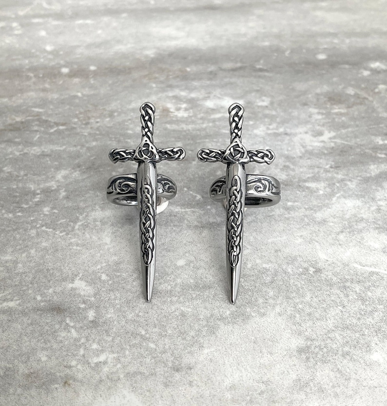 May include: Two silver rings with a sword design. The swords are detailed with intricate knotwork and have a pointed tip.